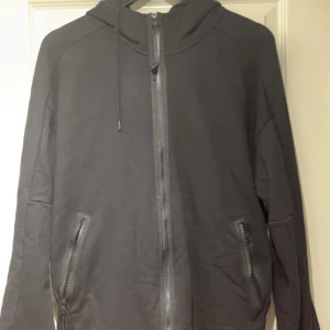 Svart hoodiejacka C.P. Company XXL - Svart hoodiejacka från C.P. Company i storlek XXL. Jackan har dragkedja framtill, två fickor med dragkedja och ikoniska goggles på huvan. Tillverkad i mjuk bomull och har en clean, urban look med långa ärmar och relaxed passform. passar L/XL