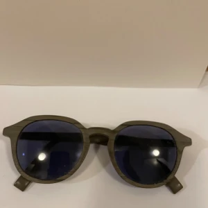 Grå runda solglasögon från HUGO - Snygga solglasögon från HUGO med rundade bågar i grått och mörka linser. Bågarna har en cool träliknande finish och logotypen HUGO på sidan. Perfekt accessoar för att lyfta din stil med en modern touch.