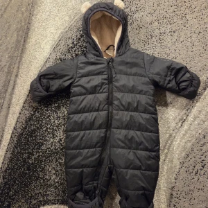 Svart fodrad overall från Lindex Baby - Mysig svart overall från Lindex Baby med quiltad utsida och fluffigt beige teddyfoder. Overall med huva och söta öron, dragkedja som går snett över framsidan och långa ärmar. Perfekt för kalla dagar!