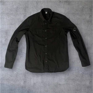 Svart Overshirt C.P. Company - Svart långärmad skjorta från C.P. Company med klassisk krage, två bröstfickor med lock och knappar hela vägen framtill. Ikonisk Stone Island-logga på vänster ärm. Skjortan har en rak passform och är tillverkad i bomull. Linsen och skjortan är som ny, bara använd under speciella tillfällen. Stryks vid beställning 🤩. Nypris på dessa varierar mellan 2500 till 4000 kr.