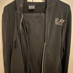 Svart zip-hoodie från EA7 Emporio Armani - Svart hoodiejacka från EA7 Emporio Armani med dragkedja och huva samt byxor till den . Jackan har snörning i huvan, en är lite slitet som på bilden,  logga på bröstet. Perfekt för en sportig och stilren look. Båda stl s