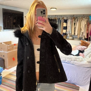 Svart rock med fuskpäls och dragkedjor - Snygg svart rock med stor huva kantad av fuskpäls. Jackan har dekorativa knappar och dragkedjor framtill samt en rak, något loose passform. Perfekt för kalla dagar när du vill vara både varm och stilren. Pälsen på luvan går att ta bort 