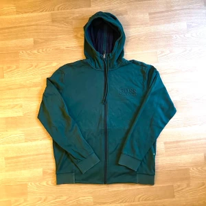 Mörkgrön hoodie från Hugo Boss - Snygg mörkgrön hoodie från Hugo Boss med dragkedja och broderad logga på bröstet. Tröjan har huva, långa ärmar och ribbade muddar. Perfekt för en chill och stilren look. Köptes för 1800kr. Säljer eftersom den inte får plats i garderoben 