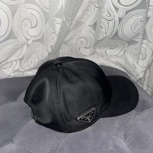Svart keps från Prada i nylon - Snygg svart keps från Prada med klassisk böjd skärm och justerbart spänne bak. Kepsen är tillverkad i glansig nylon och har Pradas ikoniska metallogo på sidan. Perfekt accessoar för en clean och lyxig streetstyle.