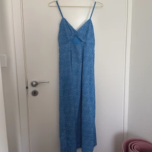 Blommig blå långklänning  - Säljer en blå långklänning med smala axelband och v-ringad front. Klänningen har ett litet cutout-detalj under bysten och har sött blommigt mönster