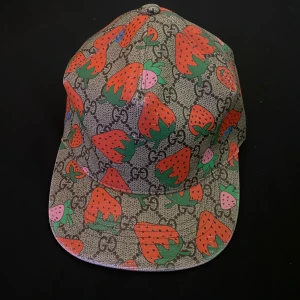 Gucci keps med jordgubbsmönster - Gucci keps med ikoniskt GG-mönster och färgglada jordgubbar i rött, rosa och grönt. Basen är beige med svarta detaljer och kepsen har justerbar rem bak. Tillverkad i bomull och polyester, fodrad med rosa tyg. Perfekt statement-piece för dig som vill sticka ut.