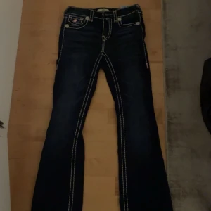 True Religion Becca Bootcut Jeans - Säljer ett par mörkblå True Religion Becca Mid Rise Bootcut jeans med tydliga kontrastsömmar och snygga fickdetaljer med knappar bak. Klassisk bootcut-modell i jeansmaterial, perfekt för dig som gillar street och statement-plagg.