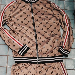 B.B. TRACKSUIT Mönstrad Khakifärgad Herr Medium - Säljer en khakifärgad tracksuit i två delar från B.B. med svart logomönster över hela, svarta och vita ribbade muddar samt röda och vita ränder längs ärmarna. Tröjan har hel dragkedja och sportig look, perfekt för dig som gillar streetwear. Utmärkt skick förutom en liten svart knappt sebar missfärgning och några noppor. Se bild 5 för inzoomning. Ej sebart utan det. 