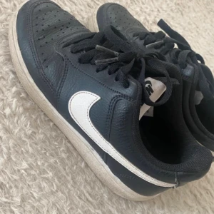 Svarta Nike sneakers med vit swoosh - Säljer ett par klassiska svarta Nike sneakers med vit swoosh på sidorna. Skorna har snörning, perforerad tå och platt sula. Tillverkade i skinn med textilfoder, perfekt för dig som gillar en clean och sportig stil.