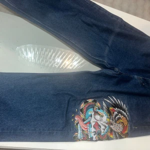 Ed Hardy jeans - Mörkblå jeans från Ed Hardy med stor vit logga på bakfickan och färgglad broderad drake på ena benet. Klassisk rak passform och cool streetstil. Perfekta för dig som vill sticka ut med detaljer och mönster.