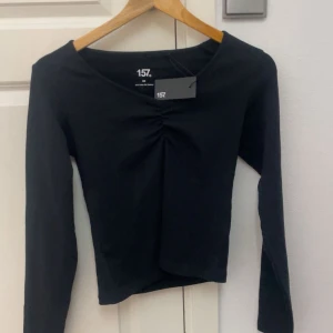 Svart långärmad topp från 157 - Svart långärmad topp från 157 med snygg rynkdetalj framtill vid bysten. Modellen är croppad och har en enkel, stilren design som passar perfekt till jeans eller kjol. Tillverkad i mjuk bomull för en skön känsla hela dagen.