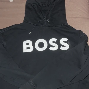 Svart hoodie från BOSS med tryck - Svart hoodie från BOSS i storlek L med stor vit BOSS-logga på bröstet. Klassisk modell med huva och snörning, ribbade muddar och mjukt material. Perfekt för en chill och stilren look.