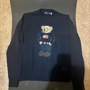 Mörkblå stickad tröja Polo Bear - Mörkblå stickad tröja från Polo Ralph Lauren med ikoniska Polo Bear broderad framtill. Tröjan har rund halsringning, ribbade muddar och RL-broderi nertill. Materialet är en mix av bomull och linne, perfekt för dig som gillar klassisk men lekfull stil.