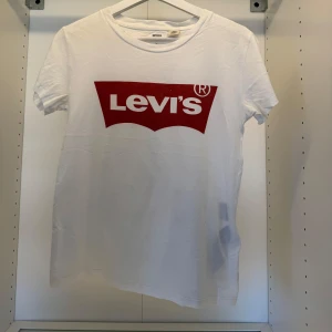 Vit Levi's t-shirt med rött tryck - Vit t-shirt från Levi's med klassiskt rött logotryck på bröstet. T-shirten har rund halsringning och korta ärmar. Tillverkad i mjuk bomull som känns skön mot huden. Perfekt för en avslappnad och stilren look.