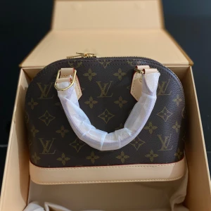 Louis Vuitton monogram handväska - Säljer en ikonisk handväska från Louis Vuitton i klassiskt monogrammönster. Väskan är brun med beiga detaljer och guldfärgade metalldelar. Tillverkad i skinn med rundad form och två handtag. Perfekt för dig som vill ha en lyxig och tidlös accessoar.