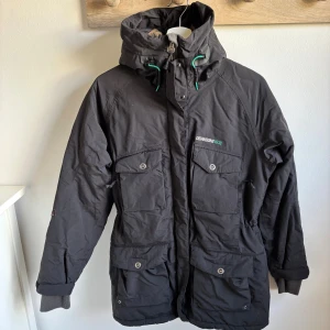 Svart parkajacka från Didriksons - Svart parkajacka från Didriksons med huva, gröna detaljer och flera stora fickor framtill. Jackan har dragkedja och knappar, samt ribbade muddar vid ärmsluten. Perfekt för kalla dagar med sin långa modell och praktiska design.