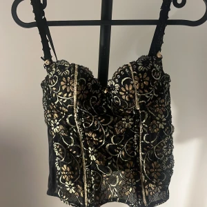 Svart och guld blommig korsettopp - Supersnygg korsettopp i svart spets med guldiga blommor och detaljer. Toppen har reglerbara axelband, markerade kupor och vackra broderade mönster framtill och baktill. Perfekt för att lyfta vilken outfit som helst med en lyxig känsla. Står att den är från märket change men de inget jag vet om:)