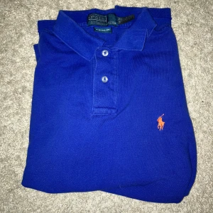 Blå Polo Ralph Lauren pikétröja M - Snygg blå pikétröja från Polo Ralph Lauren i custom fit. Tröjan har klassisk krage, två knappar och den ikoniska orangea broderade loggan på bröstet. Perfekt för dig som gillar stilrena och sportiga plagg. Materialet är mjuk bomull som andas.