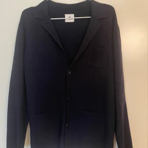 Mörkblå cardigan tröja John Henric - Snygg mörkblå cardigan jacka från John Henric med klassisk krage, tre fickor och knäppning framtill. Jackan är stickad och har långärmade ärmar, perfekt för dig som gillar stilrena och enkla plagg med lite preppy vibe.