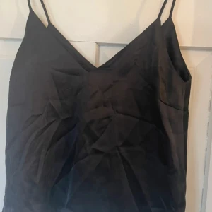 Svart linnetopp från 157 XS - Snygg svart linnetopp från 157 i storlek XS. Toppen har smala axelband och v-ringad hals, vilket ger en stilren och enkel look. Materialet har en lätt glansig finish som känns mjuk mot huden. Perfekt att styla med jeans eller kjol för en cool vibe.