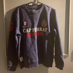 Mörkblå sweatshirt från Morris - Mörkblå sweatshirt från Morris med broderad text 'Cap Ferrat Morris' i vitt och rött framtill. Tröjan har rund hals, långa ärmar och ribbade muddar. Snygga broderade detaljer och emblem på bröstet samt tryck på ena ärmen.