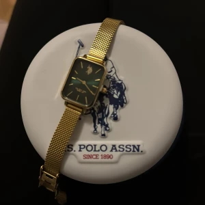 klocka från U.S. Polo Assn - Stilren klocka från U.S. Polo Assn. med rektangulärt ur och mesh-armband i guld. Urtavlan är mörkgrön med guldiga detaljer och logga upptill. 