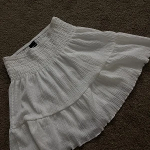 Vit volangkjol från Gina Tricot - Supersöt vit kortkjol från Gina Tricot med volang och smockad midja. Kjolen är gjord i ett lätt, krinklat bomullstyg som ger en chill och luftig känsla. Perfekt för att skapa en fräsch och trendig look med lite extra volym.