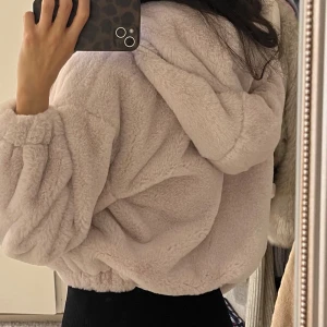 Beige teddyjacka med huva från Bershka - Supermjuk och fluffig teddyjacka från Bershka i beige färg. Jackan har oversized passform, huva med snörning och dragkedja framtill. Perfekt för kalla dagar och ger en chill vibe. Resår vid ärmslut och midja för extra komfort.