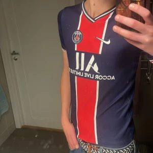 PSG fotbollströja Nike Dri-Fit L - Säljer en snygg Paris Saint-Germain fotbollströja från Nike i storlek L. Tröjan är marinblå med en bred röd rand och vita detaljer, har korta ärmar och V-ringad krage. Materialet är Dri-Fit polyester som andas och håller dig torr under matchen.