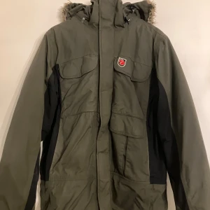 Grön parkajacka från Fjällräven S - Parkajacka från Fjällräven i mörkgrön och svart färg med huva och fuskpäls. Jackan har flera stora fickor framtill, dragkedja och knappar. Materialet är polyamid och polyester, perfekt för kalla dagar. Klassisk Fjällräven-logga på bröstet.