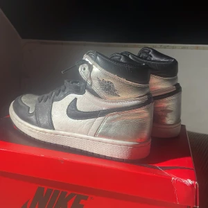 Nike Air Jordan 1 Silver/Black - Säljer ett par Nike Air Jordan 1 i en riktigt clean färgway med silver och svart. Skorna har klassisk hög siluett, svart swoosh och detaljer i skinn. Silvermetallic paneler ger en unik vibe och de har svart snörning samt svart yttersula. Perfekta för dig som vill sticka ut med dina sneakers.