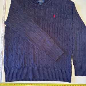 Mörkblå kabelstickad tröja Polo Ralph Lauren - Kabelstickad mörkblå tröja från Polo Ralph Lauren med klassisk rund halsringning och röd broderad logga på bröstet. Tröjan har långa ärmar och breda ribbade muddar vid ärmslut och nederkant. Perfekt för lager-på-lager under kyliga dagar. Denna kofta är i L barnstorlek eller 15-16 år. Du hittar mått på bilden