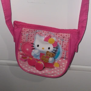 Hello Kitty rosa axelväska - Söt rosa axelväska med Hello Kitty-tryck på framsidan. Väskan har ett färgglatt motiv med prickigt och blommigt mönster, samt en lång axelrem. Perfekt för dig som gillar gulliga accessoarer och vill ha något unikt till din outfit.