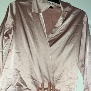 Champagnefärgad omlottblus från Bik Bok - Snygg ljusrosa omlottblus från Bik Bok i glansigt satinmaterial. Blusen har långa ärmar, klassisk krage och knytband i midjan för en cool look. Perfekt för dig som gillar stilrena och trendiga plagg med lite extra shine.