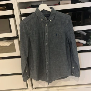 Blå skjorta från Ralph Lauren L 14-16 - Snygg blå skjorta från Ralph Lauren med klassisk button-down krage och broderad logga på bröstet. Skjortan har lång ärm, knappar framtill och är tillverkad i mjuk bomull. Perfekt för dig som gillar stilrena och tidlösa plagg.