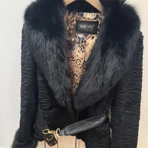 Svart äkta pälsjacka från Hollies - Säljer en svart pälsjacka från Hollies med bred, fluffig krage och snyggt bälte med gulddetaljer. Jackan har ett coolt leopardmönstrat innerfoder och markerad struktur i materialet. Perfekt för dig som vill sticka ut med en lyxig vibe.