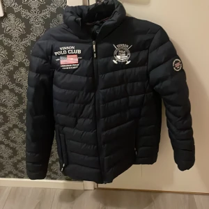 Navy puffer jacket - Snygg mörkblå pufferjacket från Vinson Polo Club, loggor med amerikansk flagga på bröstet och ärmen. Jackan har ståkrage, dragkedja och quiltad design som ger en sportig vibe. Perfekt för kalla dagar. Helt i nyskick!!! Använd Max 2-3 gånger. Säljes pga den är för liten