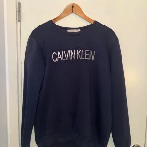 Marinblå sweatshirt från Calvin Klein - Snygg marinblå sweatshirt från Calvin Klein med stort logotryck i silver på bröstet. Tröjan har rund hals och långa ärmar, perfekt för en chill och stilren look. Mjuk bomullskänsla och klassisk passform.