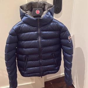 Mörkblå Colmar pufferjacka med huva - Snygg mörkblå pufferjacka från Colmar med huva och dragkedja framtill. Jackan har quiltad design, två fickor med dragkedja och Colmar-logga på ärmen. Insidan är grå och materialet känns lätt men ändå varmt, perfekt för kyliga dagar.