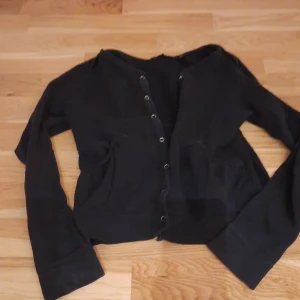 Svart kofta med knappar - Svart kofta med långa ärmar och knäppning framtill. Tröjan har två fickor på sidorna och är i en enkel, klassisk modell. Perfekt för lager-på-lager och passar till det mesta. Storlek 146/152.