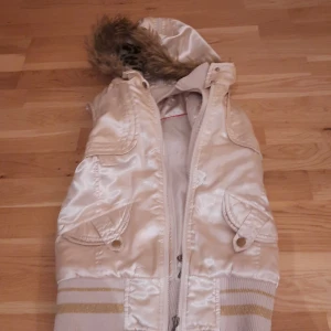 Beige vintage dunväst med leopardfoder och huva - Snygg beige dunväst med glansig yta, huva med fuskpäls och leopardmönstrat foder. Västen har dragkedja framtill, två fickor med lock och ribbade muddar med guldiga ränder. Perfekt för dig som vill sticka ut med coola detaljer och djurmönster.