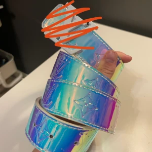 Holografiskt bälte med silverdetaljer - Säljer ett coolt holografiskt bälte med stora silverdetaljer och spänne. Bältet har en skimrande yta i regnbågsfärger och är perfekt för att ge outfiten en unik touch.