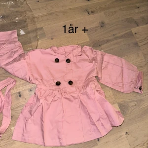 Rosa trenchcoat för barn med bälte kappa - Söt rosa trenchcoat för barn med dubbelknäppning framtill och tillhörande bälte. Jackan har lång ärm, veckad kjoldel och stora knappar i mörk kontrast. Perfekt för dig som vill ha en gullig och trendig jacka till de små.Söt rosa kappa för barn, perfekt för en liten fashionista. Kappan har en klassisk design med knappar fram och ett fint skärp i midjan. Den utställda kjolen ger en lekfull touch.
