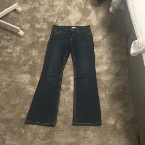 Mörkblå bootcut jeans Gina Tricot - Snygga mörkblå jeans från Gina Tricot i bootcut-modell med kontrasterande vita sömmar. Klassiska femficksjeans med lock på bakfickorna och silverfärgade knappar. Perfekta för dig som gillar en retro vibe och vill ha ett par jeans med lite extra attityd.