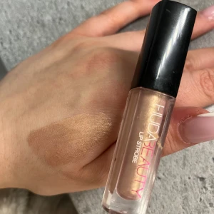Mini Huda Beauty Lip Strobe i guld - Glansigt läppglans från Huda Beauty i nyansen Lip Strobe, med skimrande guldton och metallic finish. Kommer i en smidig tub med svart lock och applikator. Perfekt för att ge läpparna extra glow och volym. Ungefär mer än halv kvar 