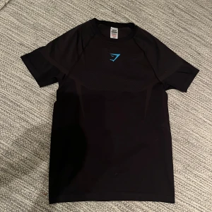 Gymshark onyx 5.0 - Svart tight t-shirt från Gymshark med blå logga på bröstet. T-shirten har korta ärmar, sportig passform och meshdetaljer upptill för extra ventilation. Perfekt för gymmet eller annan träning. Materialet är stretchigt och andas bra.