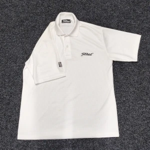 Vit Titleist pikétröja i polyester - Fräsch vit pikétröja från Titleist med diskret rutmönster i tyget och broderad logga på bröstet. Tröjan har klassisk krage, korta ärmar och knappar framtill. Perfekt för golf eller chill dagar. Materialet är lätt polyester som andas.