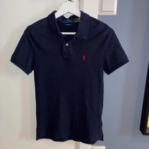 Mörkblå Polo Ralph Lauren piké - Snygg mörkblå piké från Polo Ralph Lauren med klassisk krage och två knappar framtill. Röda loggan broderad på bröstet. 
