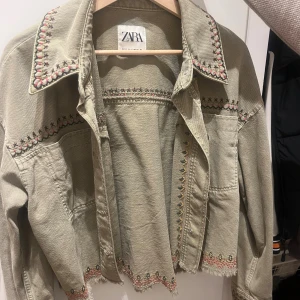 Ljusgrön broderad jeansjacka från Zara - Snygg ljusgrön jeansjacka från Zara med broderade detaljer i rosa, svart och grönt längs krage, axlar och nederkant. Jackan är croppad med fransad kant och har långa ärmar. Kan diskutera pris!