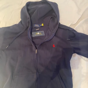 Marinblå hoodie från Polo Ralph Lauren - Snygg marinblå hoodie från Polo Ralph Lauren med dragkedja och huva. Tröjan har ett rött broderat Polo-logo på bröstet, snörning vid huvan och praktiska fickor framtill. Tillverkad i mjuk bomull för skön komfort.